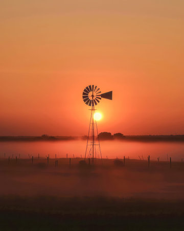 Silhouette of a windmill at dawn with mist and a vibrant orange sunrise in the background.