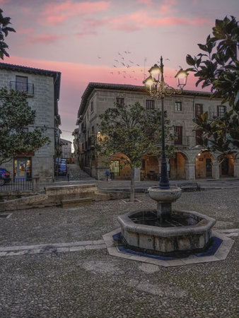 The square from Spain in Briones, La Rioja, Spain, featuring a stone fountain, classic streetlights, and traditional architecture under a colorful sunset sky.の写真素材