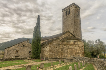 Romanesque stone church with a prominent bell tower and small cemetery, set in a peaceful rural landscape with mountain views.の写真素材