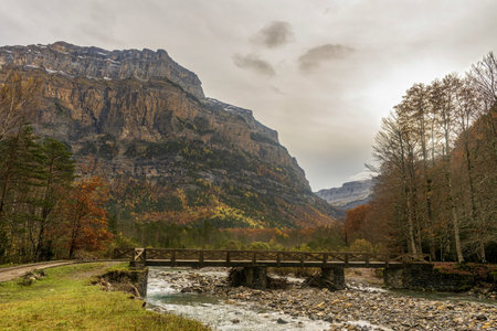 Rustic wooden bridge crossing a clear river in a dramatic valley, surrounded by cliffs and vibrant forest scenery.の写真素材