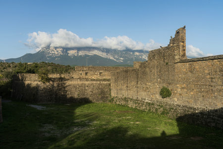 Ruins of a stone fortress with lush green surroundings and a stunning mountain range in the background under a clear blue sky.の写真素材