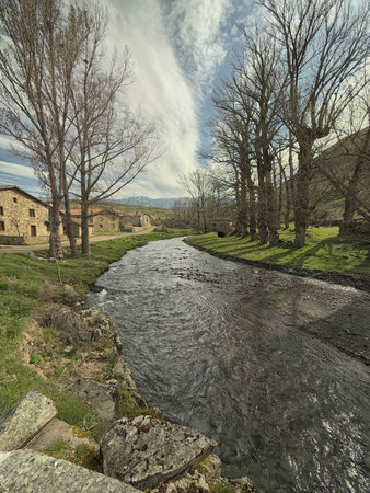 Peaceful riverside landscape with stone houses, trees, and Pisuerga River in San Juan de Redondo, Spain.の写真素材