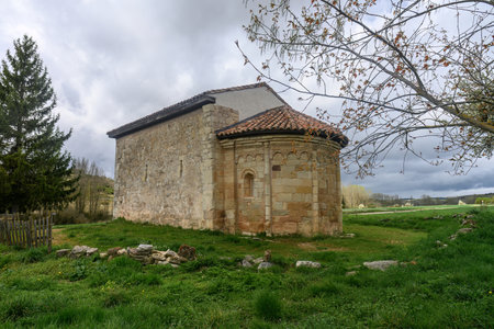 Romanesque hermitage with stone walls and apse, located in a green rural landscape under cloudy skies in Spain.の写真素材