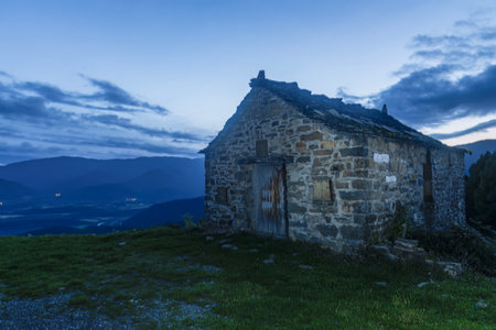 A solitary stone cabin with weathered walls sits on a grassy hilltop, overlooking a peaceful mountain valley during twilight.の写真素材