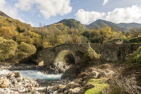 Ancient stone bridge crossing a flowing river, nestled in a picturesque valley with vibrant foliage and mountains.の写真素材