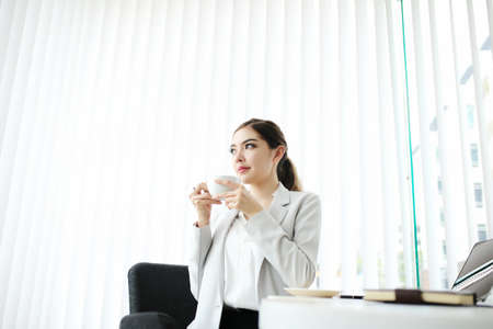 Young pretty woman sitting at opened window looking outside enjoys of rest. charming businesswoman with suit hand hold hot coffee cup drink in morning. Good life feeling roasted bean aromaの写真素材