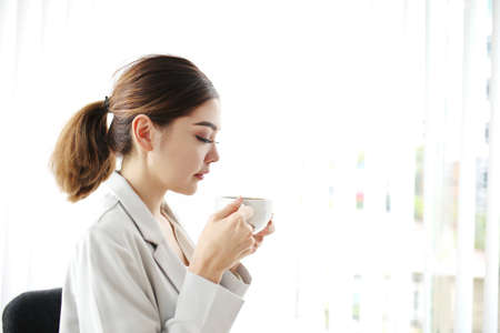 Young pretty woman sitting at opened window looking outside enjoys of rest. charming businesswoman with suit hand hold hot coffee cup drink in morning. Good life feeling roasted bean aromaの写真素材