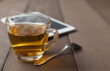 Close up hot tea cup on wood table with tea spoon and tablet shot on moning time witn sun ray background. Soft focus and selective focus to tea cup. hot tea in glass with copy space for text or design.の写真素材