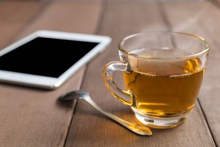Close up hot tea cup on wood table with tea spoon and tablet shot on morning time with sun ray background. Soft focus and selective focus to tea cup. hot tea in glass with copy space for text or design.の写真素材