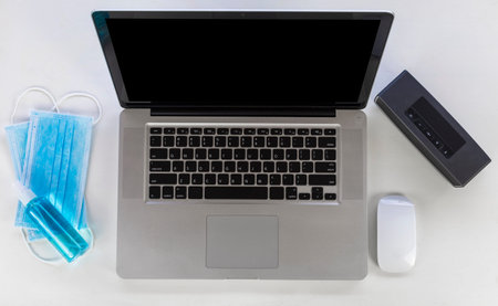 Top view Laptop, mouse, speaker, alcohols spray and Medical mask on white wood table. Work from home...