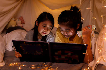 Family concept. elder sister and sister reading book with flashlight together before bedtime. Sister read story book together in bed sheet tent. focus selective little sister. With film grain effectの写真素材