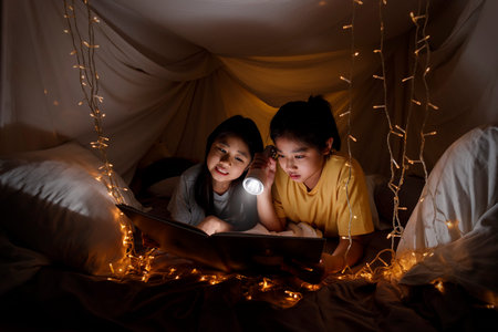 Family concept. elder sister and sister reading book with flashlight together before bedtime. Sister...