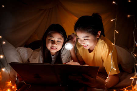 Family concept. elder sister and sister reading book with flashlight together before bedtime. Sister read story book together in bed sheet tent. focus selective little sister. With film grain effectの写真素材