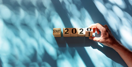 Hand holding wooden cube with flip over block start 2020 to 2021 word on blue and shadow background. Top view hand flip wood block with number text start 2020, 2021 with copy spaceの写真素材