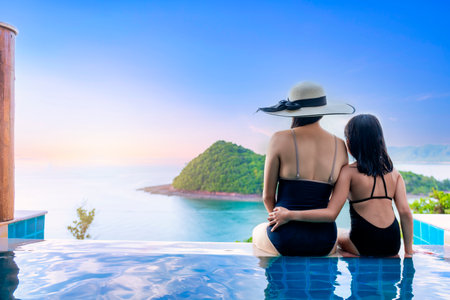 Asian women in black swimsuit with her daughter siting on side of infinity pool relaxing at sunset time  with sea view during vacation retreat. Family happy and enjoy with holiday summer travel time.の写真素材