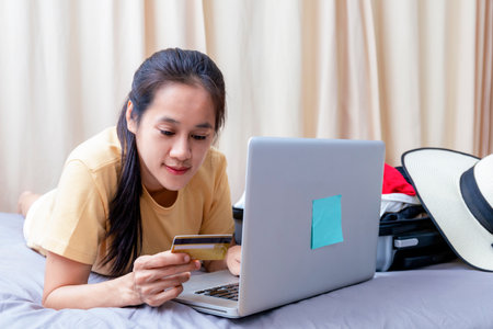 Asian happy woman plan for trip by laptop and credit card on the bed at home. Women with travel bag use notebook computer and credit card pay booking hotel. Holiday summer conceptの写真素材