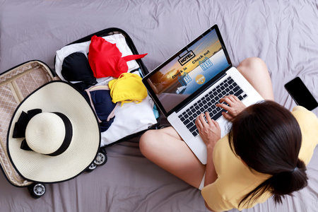 Top view Asian happy woman plan for trip by laptop booking hotel on the bed at home. Women with travel bag use notebook computer internet booking hotel. Holiday summer vacation conceptの写真素材