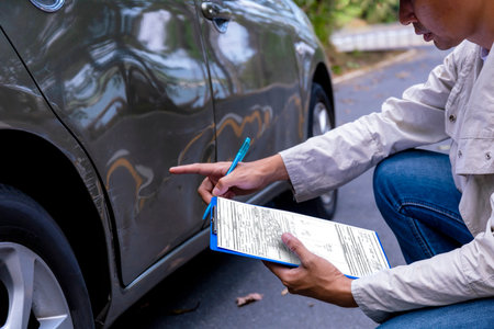 Insurance agent writing on clipboard while examining car after accident claim being assessed and...