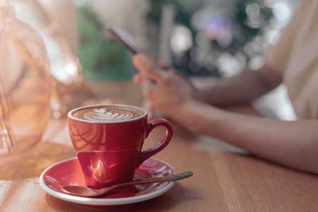 Close up hot cappuccino, Latte coffee with blur hands of woman holding mobile, smart phone sitting...