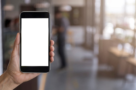Hand man holding black mobile phone with blank white screen with blur coffee shop background. Mockup...