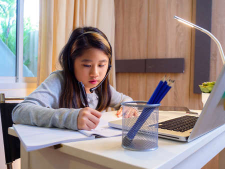 Asian girl in headphones sit at desk study online on laptop. Kid wear headset handwriting in notebook learning using internet lessons on quarantine. Girl student learning virtual internet online class from school due to pandemic.の写真素材