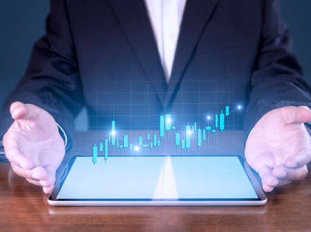 Hand of businessman present tablet analysis stock market graph growth and increase of chart positive indicators. Hand holding tablet show stock chart on blue background.の写真素材