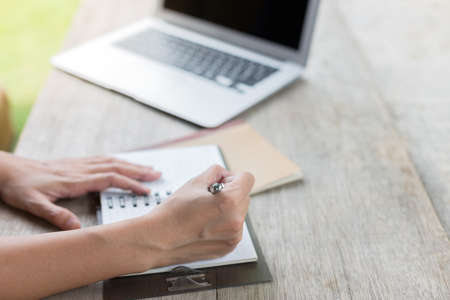 A woman holding a pen is writing on a notebook with a laptop and a coffee mug on a wooden table with warm lightの写真素材