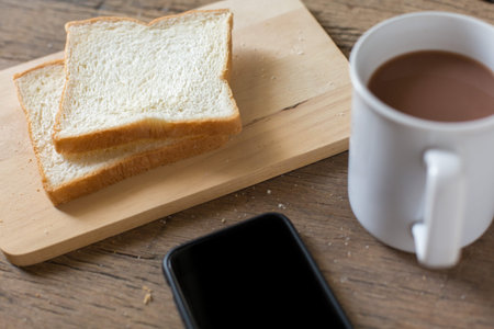 Breakfast bread and coffee in a white cup and Smart phone on a wooden table with morning sunlight...