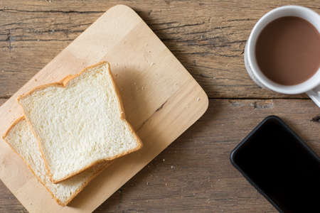 Top view of Breakfast bread and coffee in a white cup and Smart phone on a wooden table with morning sunlight and copy spaceの写真素材