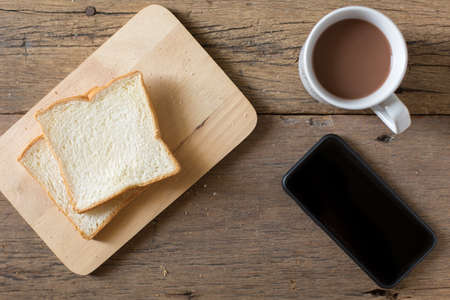 Top view of Breakfast bread and coffee in a white cup and Smart phone on a wooden table with morning sunlight and copy spaceの写真素材