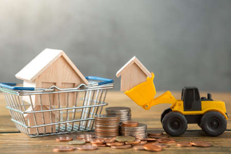 Model house in shopping basket, loader, coins stack on wooden desks and loft walls with sunlight and copy space for money saving conceptの写真素材