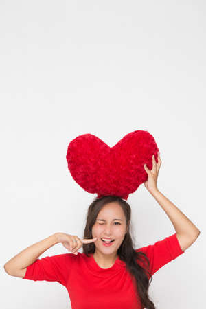 Beautiful Asian woman wearing small Santa hat and wearing red dress holding red heart shape pillow on white background. Cute Asian woman smiling and cheerful on Christmas and Valentine Day.の写真素材