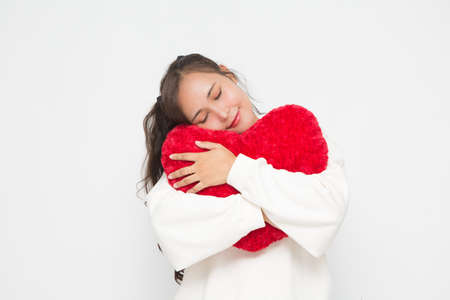 Beautiful Asian woman wearing white sweater closing her eyes and hugging red heart shape pillow on white background and copy space. Cute Asian woman smiling and cheerful for Valentine Day concept.の写真素材