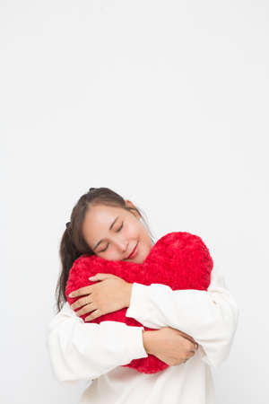 Beautiful Asian woman wearing white sweater closing her eyes and hugging red heart shape pillow on white background and copy space. Cute Asian woman smiling and cheerful for Valentine Day concept.の写真素材