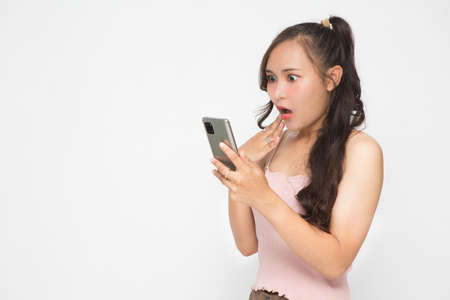 Beautiful Asian woman wearing pink casual shirt holding smartphone on white background and copy space. Cute Asian woman is shocked something on her smartphone for social media conceptの写真素材