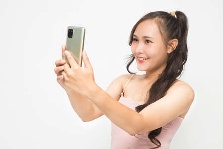 Beautiful Asian woman wearing pink casual shirt holding smartphone and selfie on white background and copy space. Cute Asian woman smiling and cheerful having fun selfies for social media conceptの写真素材