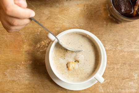 Hand holding spoon scooping Creamy mushroom soup in white cup on the wood table in restaurant.の写真素材