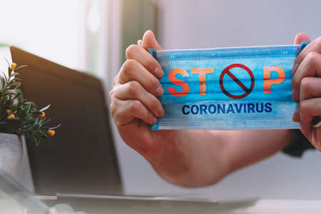 The hand was holding a mask, with the words stop coronavirus. There was a laptop in the office at home. Concept of stopping covid-19 infection for the nation and at homeの写真素材