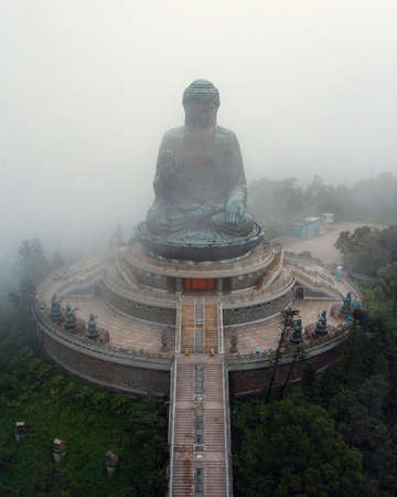 big buddha statue on top of a large mountain with fog surrounding it in backgroundの写真素材