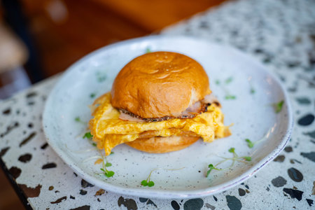 delicious egg sandwich on brioche bun with ham cheese and scrambled egg garnished with small green leaves at brunch restaurantの写真素材