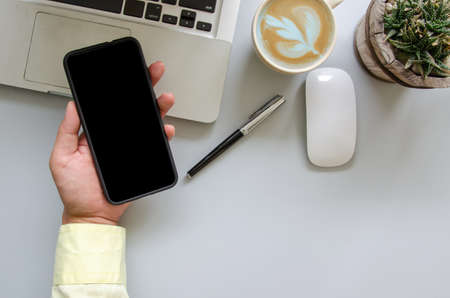 Male hand holding smartphone ,computer ,pen, coffee, mouse  on office desk table with copy spaceの写真素材