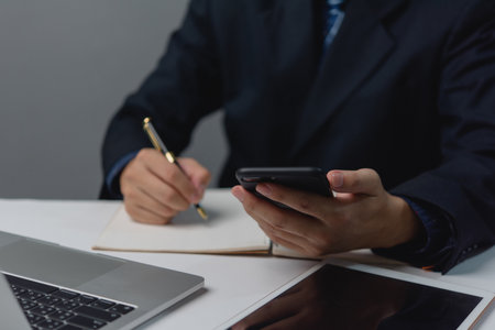 A man in a suit is writing on a piece of paper with a pen while holding a cell phone. The scene suggests a professional setting, possibly in an office or a meeting room. The man's focus on the paperの写真素材