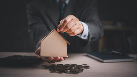 Hand placing coin into wooden house model, symbolizing savings and investment. Ideal for financial planning, real estate, mortgage, banking. Concept of economic growth and secure investments.の写真素材