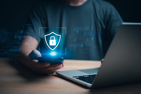 Hand holding smartphone with cybersecurity hologram, laptop in background. Represents data...