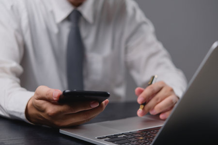 Businessman holding smartphone, using laptop. Managing tasks, modern office setup. Focus on digital...