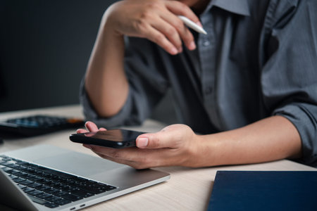 Person holding a smartphone while working on a laptop, symbolizing multitasking and modern business...