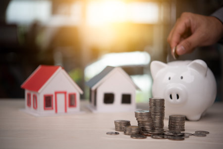 Hand placing coin into piggy bank with house models and stacked coins, representing savings and property investment. Real estate, finance, and homeownership concepts. Finance and real estate industryの写真素材
