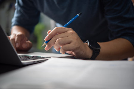 Person holding pencil while typing on a laptop, focused on work. Suitable for corporate,...