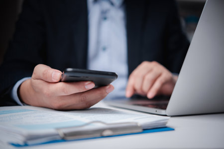 A close-up shot of a businessman holding a smartphone and typing on a laptop, focused on business work.の写真素材