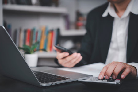 Businessman using calculator and smartphone at desk with laptop, symbolizing financial planning,...